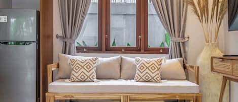 A wooden sofa with patterned cushions, a window with curtains, a refrigerator, and a vase with dried plants.