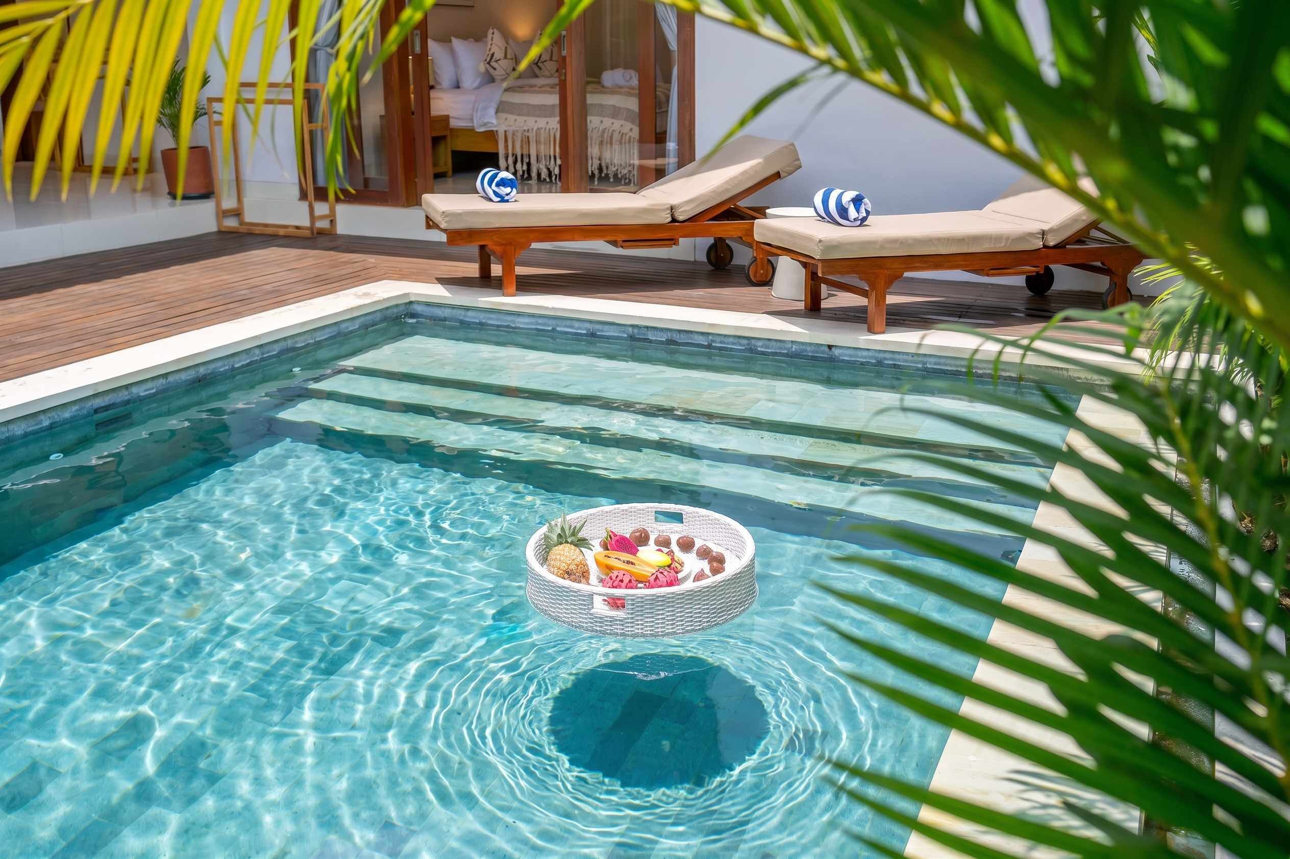 Piscina cristalina com espreguiçadeiras e uma cesta de frutas flutuando no centro.