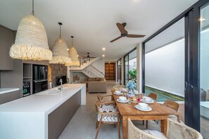 A modern kitchen with a central island, dining area with wooden table and chairs, and a view of a pool through large windows. - Brand NEW Villa+Amazing Jungle View+Huge Pool+4BDR (Kabupaten Badung)