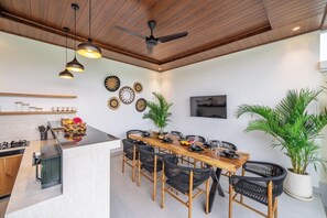 A modern kitchen with a wooden ceiling, a dining area with black wicker chairs, and a large potted plant. - Brand NEW Villa+Rice Fields View+Huge Pool+4BDR (Kecamatan Kuta Utara)