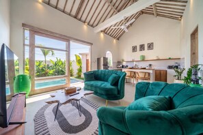 A modern living space with a large sliding glass door leading to a pool, two green sofas, a flat-screen TV, and a kitchen area with a dining table. - Brand New Paradise 2BDR Villa, Large Pool, Canggu (Kabupaten Badung)
