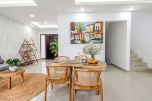 A modern living room with a wooden dining table, wicker chairs, a painting of a canal scene, and a staircase leading to another level. - Chic 3BR in Nusa Dua - Ocean View, Cinema, Rooftop (Kabupaten Badung)