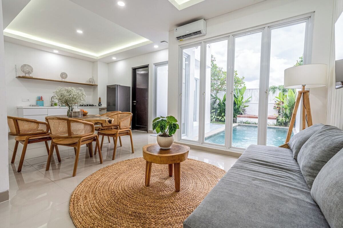 Una sala moderna con un sofá, un comedor con sillas, una mesa de centro con una planta y vistas a una piscina a través de amplios ventanales.