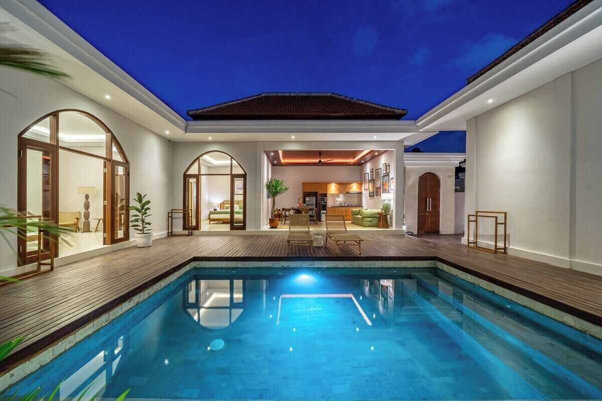 A modern indoor swimming pool with a wooden deck, a dining area, and a kitchen visible through an archway.