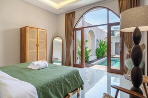 A hotel room with a bed, a wooden wardrobe, a mirror, a lamp, and a view of a swimming pool and outdoor area. - Brand NEW Paradise Villa, Huge Pool, Best Location (Kabupaten Badung)