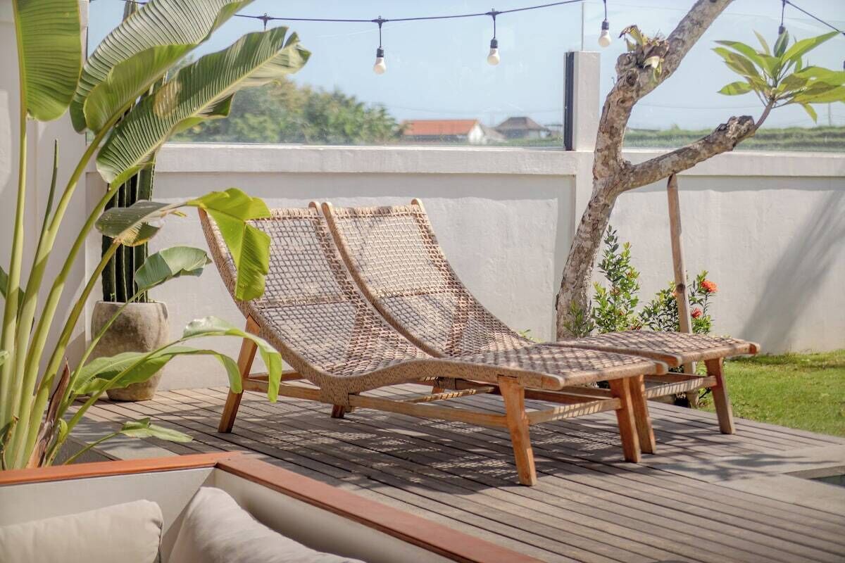 A deck with two wicker lounge chairs and a small table, surrounded by potted plants and a tree.