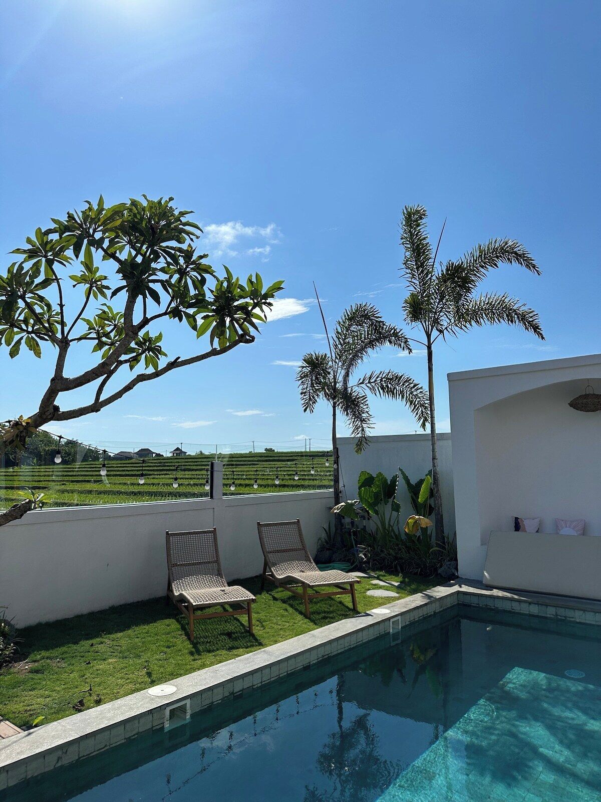 A swimming pool with two lounge chairs, a white wall, and a view of a green field.