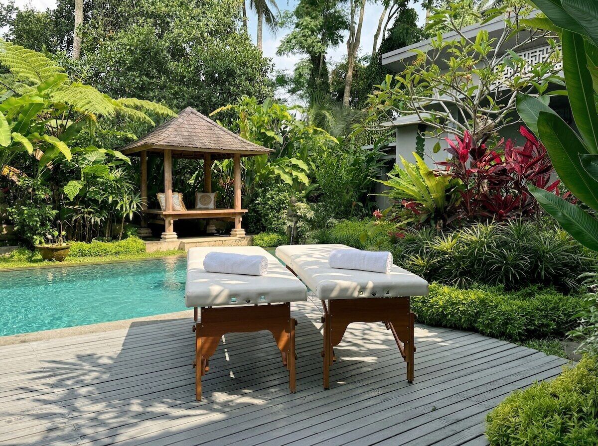 Um deck de madeira com duas mesas de massagem, um gazebo e uma piscina cercada por vegetação exuberante.