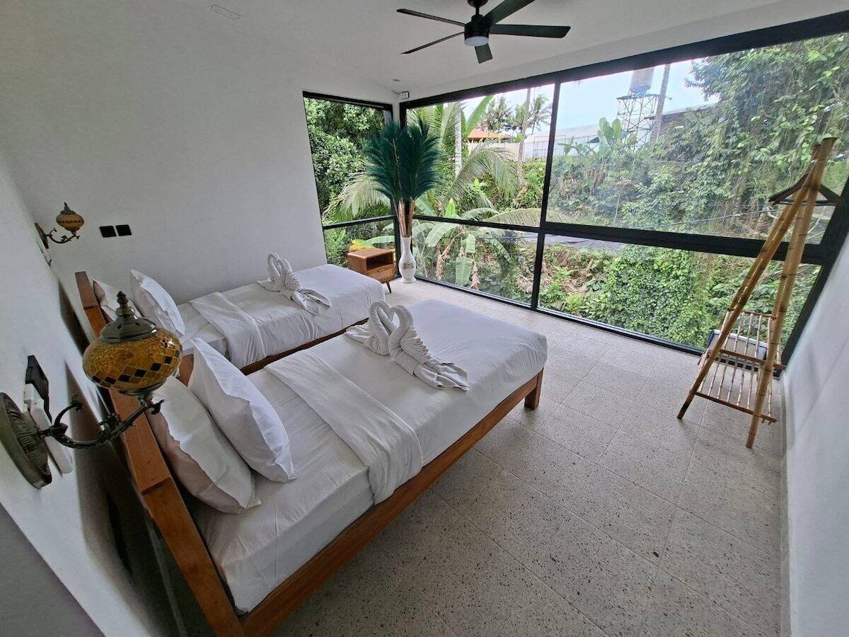 A bedroom with a large bed, a ceiling fan, a window with a view of greenery, and a small table with a plant.