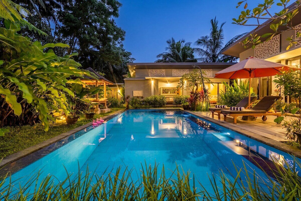A swimming pool with lounge chairs and umbrellas, surrounded by tropical plants and a building with decorative wall panels.