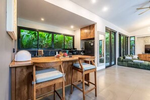 A modern kitchen with a bar area, wooden stools, and a large window with a view of greenery. - Tropical 4BR Oasis w Huge Pool+Gazebo, Ubud's Best (Kabupaten Gianyar)