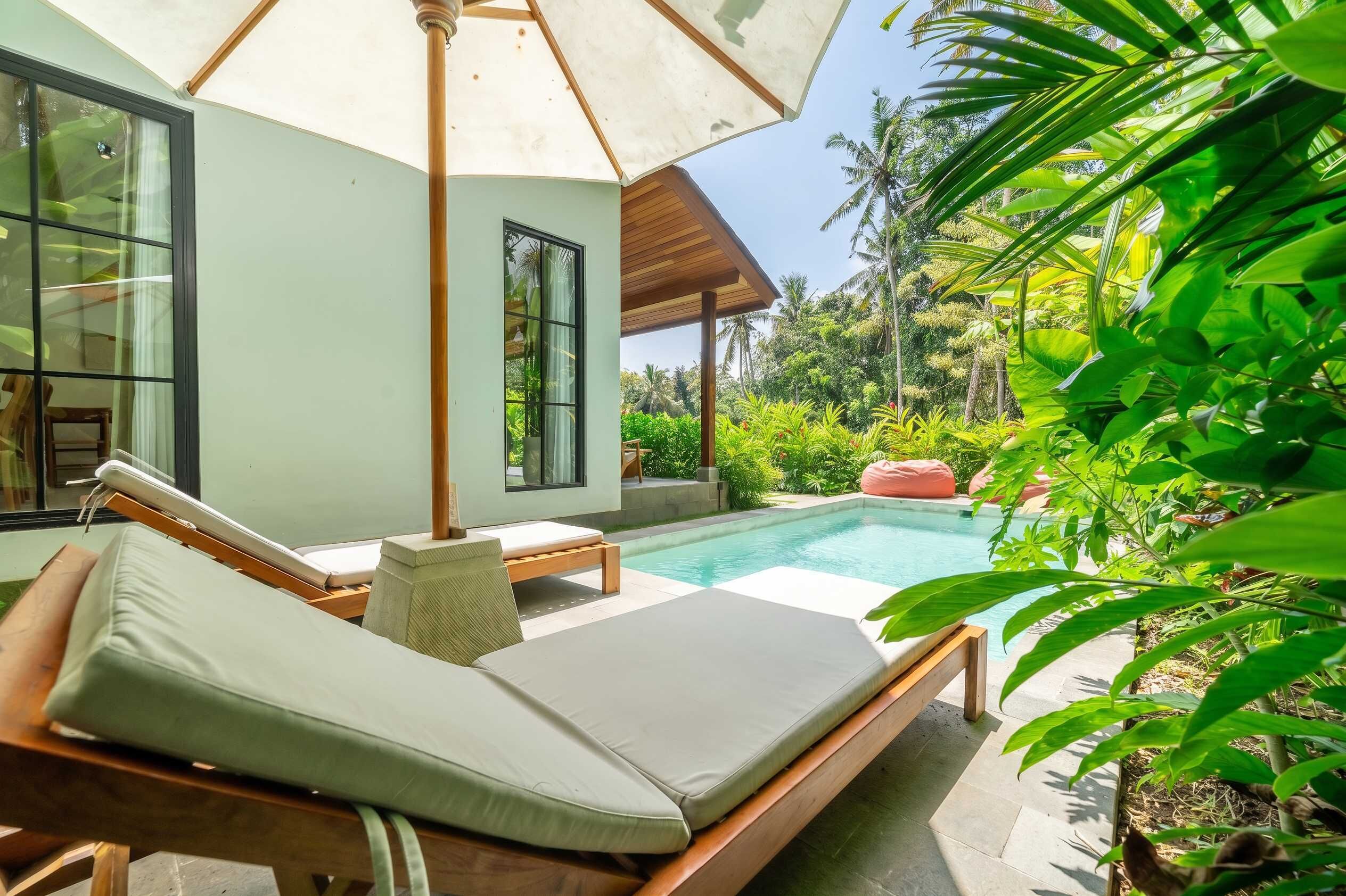 Un espace de détente aménagé au bord de la piscine, avec un banc en bois et un fauteuil confortable, une pièce fermée par des baies vitrées et une vue sur un jardin tropical.