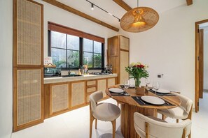 A modern kitchen with a dining area, featuring a large window, wooden cabinetry, and a dining table set for four. - Serene 2BR Stay in Top Ubud's Spot w Pool & Garden (Kabupaten Gianyar)