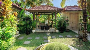 A garden with a pergola, potted plants, a stone pathway, and a building with a tiled roof. - Peaceful Ubud's 1BR • Lush Garden • Pool • Patio (Gianyar Regency)
