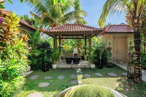 Ein Garten mit einer Pergola, Topfpflanzen, einem Kiesweg und einem Gebäude mit einem Ziegeldach.
