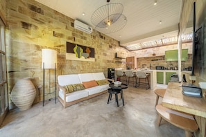 A modern living room with a stone wall, a white sofa, a coffee table, and a dining area with a wooden table. - Serene Ubud 2BR Stay • Garden Bath • Private Pool (Ubud)