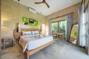 Een slaapkamer met een groot bed, een plafondventilator, een raam met gordijnen en uitzicht op groen.