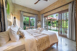 Un dormitorio amplio con una cama grande, un ventilador de techo y vistas a un jardín a través de ventanas amplias.