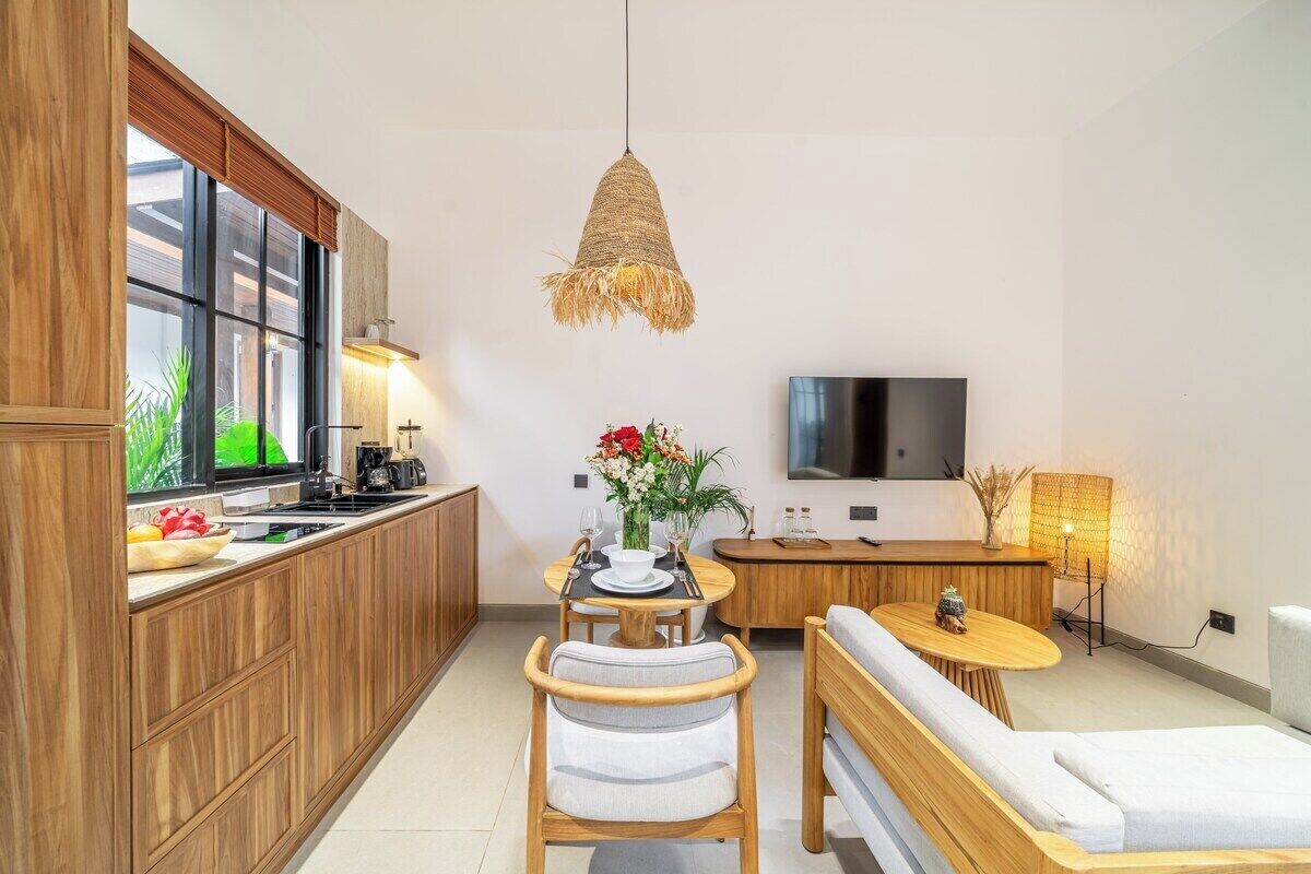 A compact kitchen with wooden cabinets, a dining area with a table and chairs, a sofa, and a TV mounted on the wall.