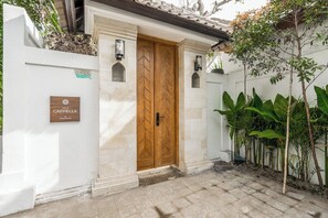 A villa entrance with a wooden double door, a white wall with a plaque, and a small garden area with plants. - Peaceful 3BR Villa in Lush Ubud, Large Pool&Garden (Kabupaten Gianyar)