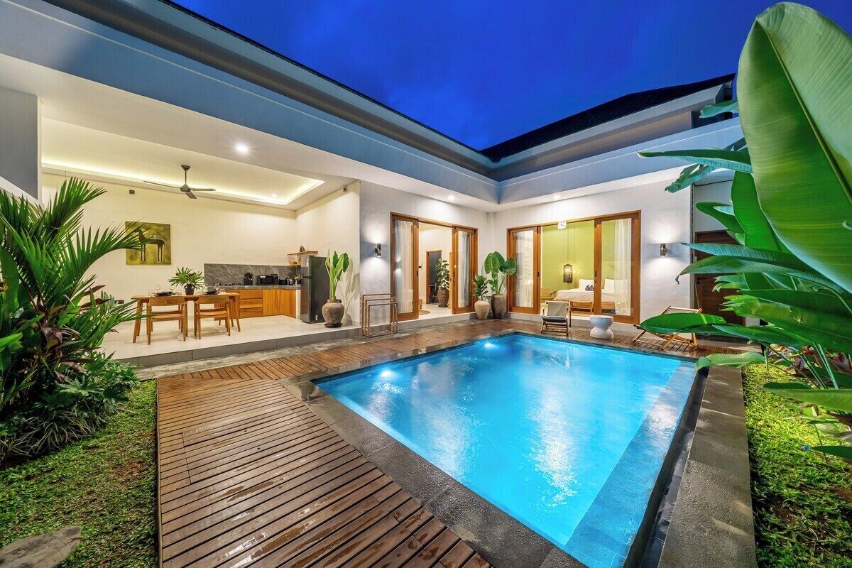 Piscina moderna ao ar livre com deck de madeira, cercada por vegetação exuberante e um espaço interior bem iluminado com área de jantar.