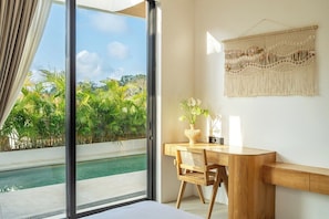 A room with a large sliding glass door leading to a pool, a wooden desk with a chair, and a macrame wall hanging. - New 1BR Villa+Beach Access+Large Pool+South Kuta (Badung Regency)
