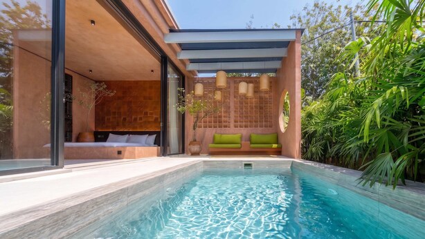 Un área de piscina moderna al aire libre con una piscina azul cristalina, una zona de estar con cojines y una pérgola de madera con luces colgantes.