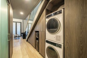 Une buanderie équipée d’une machine à laver et d’un sèche-linge superposés, avec un plancher en bois et un escalier menant à l’étage supérieur.