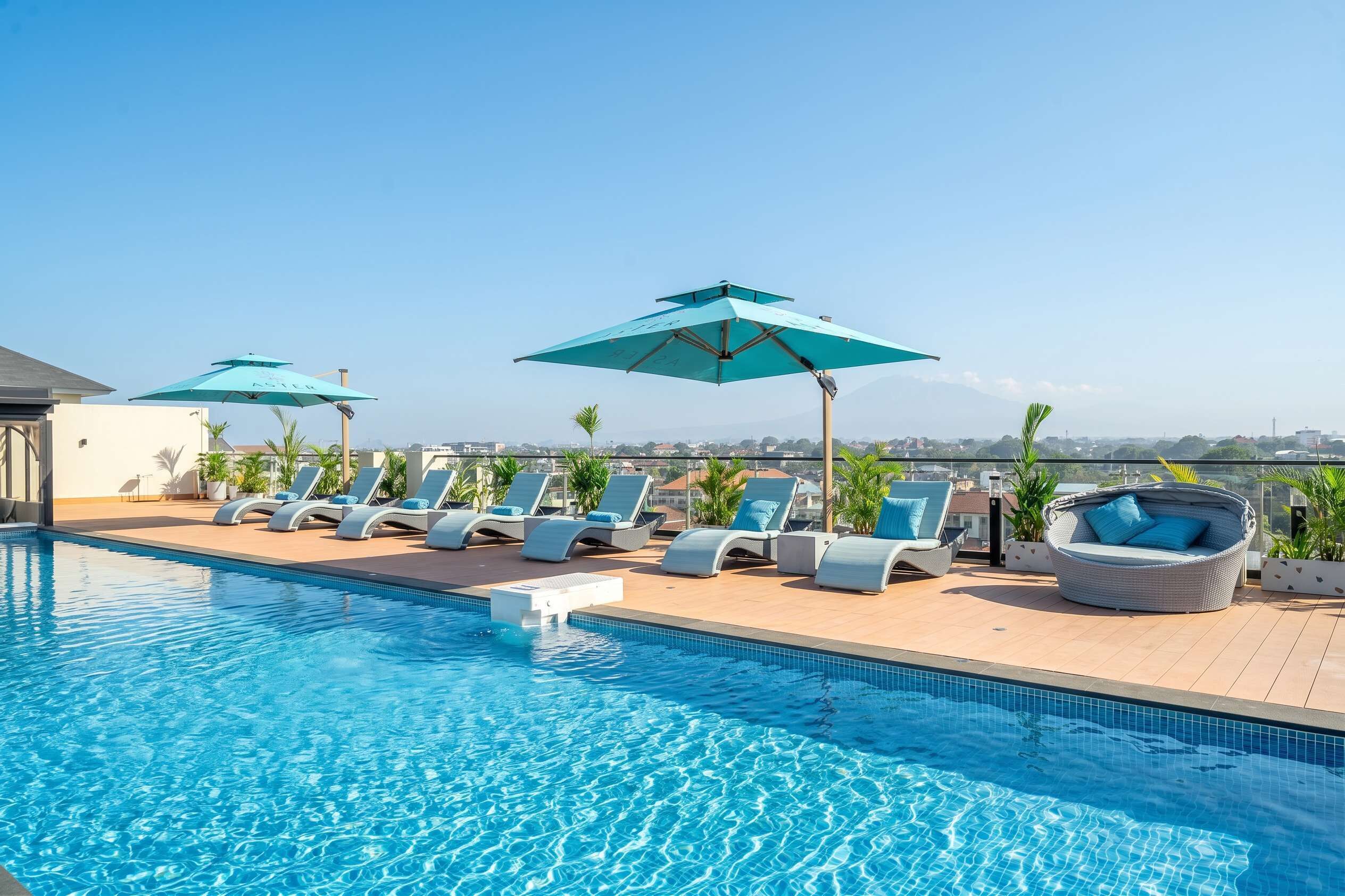 Un área de piscina en la azotea con sillones de descanso y sombrillas, cielo despejado y vistas de la ciudad.