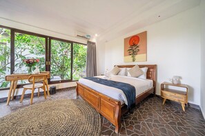 A bedroom with a bed, a bedside table, a chair, a window with a view of greenery, and a painting on the wall. - Cozy 1BR Villa in Lush Ubud w Spa+2 Pools+Balcony (Ubud)
