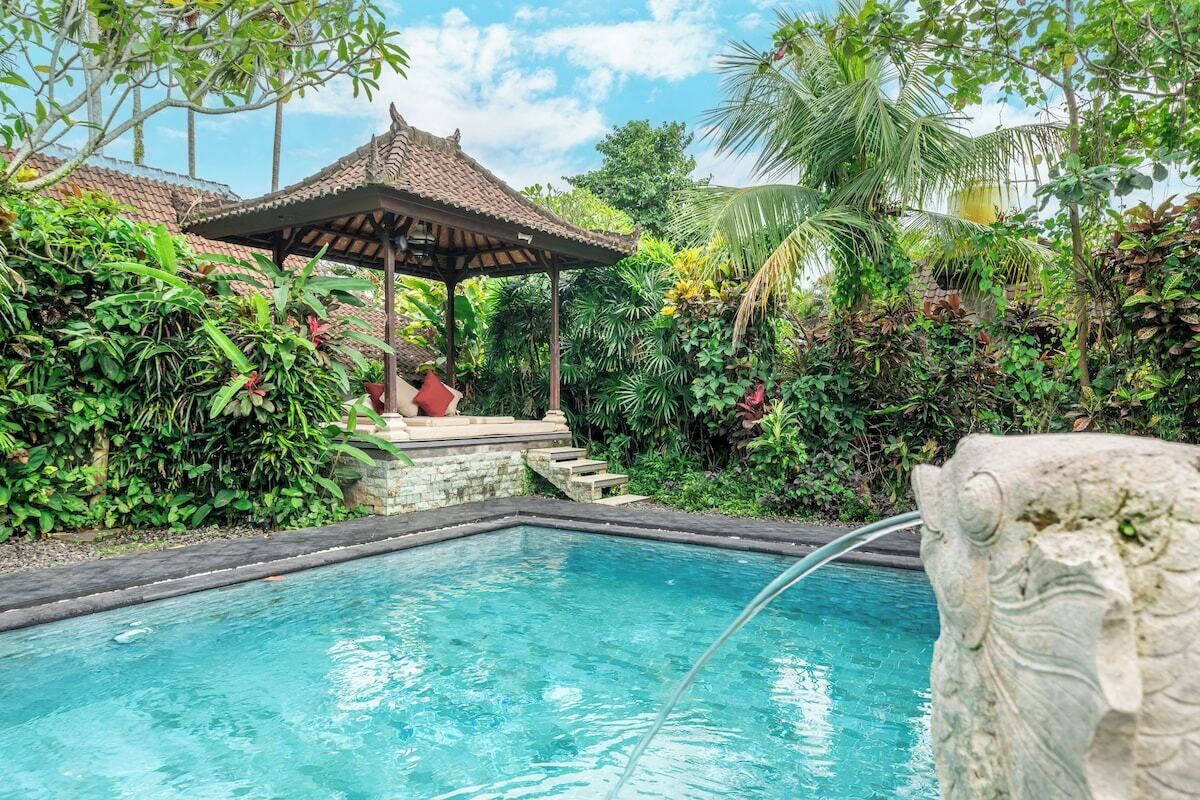 Une piscine agrémentée d’une sculpture en pierre, entourée d’une végétation luxuriante et d’un belvédère de style traditionnel.