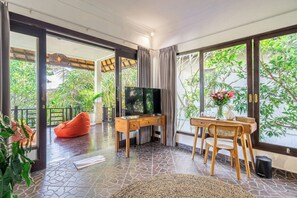 A room with a TV on a wooden desk, a red bean bag, a dining table with chairs, and a view of greenery through large windows. - Cozy 1BR Villa in Lush Ubud w Spa+2 Pools+Balcony (Ubud)