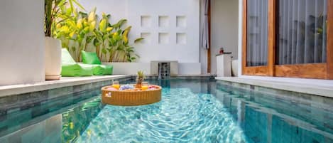 A clear swimming pool with a floating fruit basket, surrounded by potted plants and a modern building with large windows.