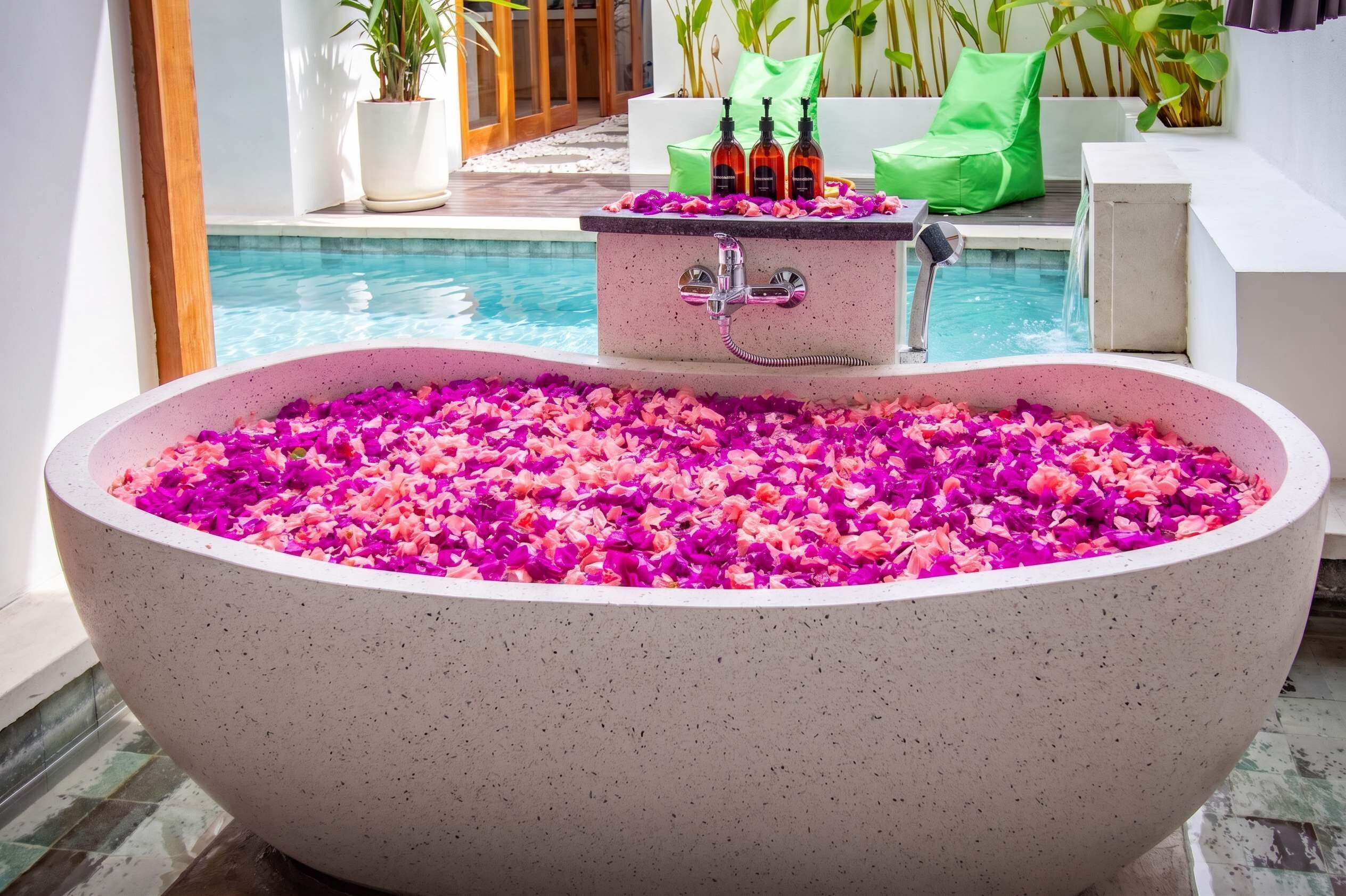 Eine große Badewanne voller Blütenblätter, ein kleines Waschbecken mit Flaschen und ein Pool im Hintergrund.