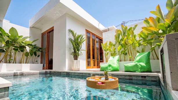 Un espace piscine moderne avec une piscine à l’eau d’un bleu limpide, un panier en osier rempli de boissons et des coussins verts.
