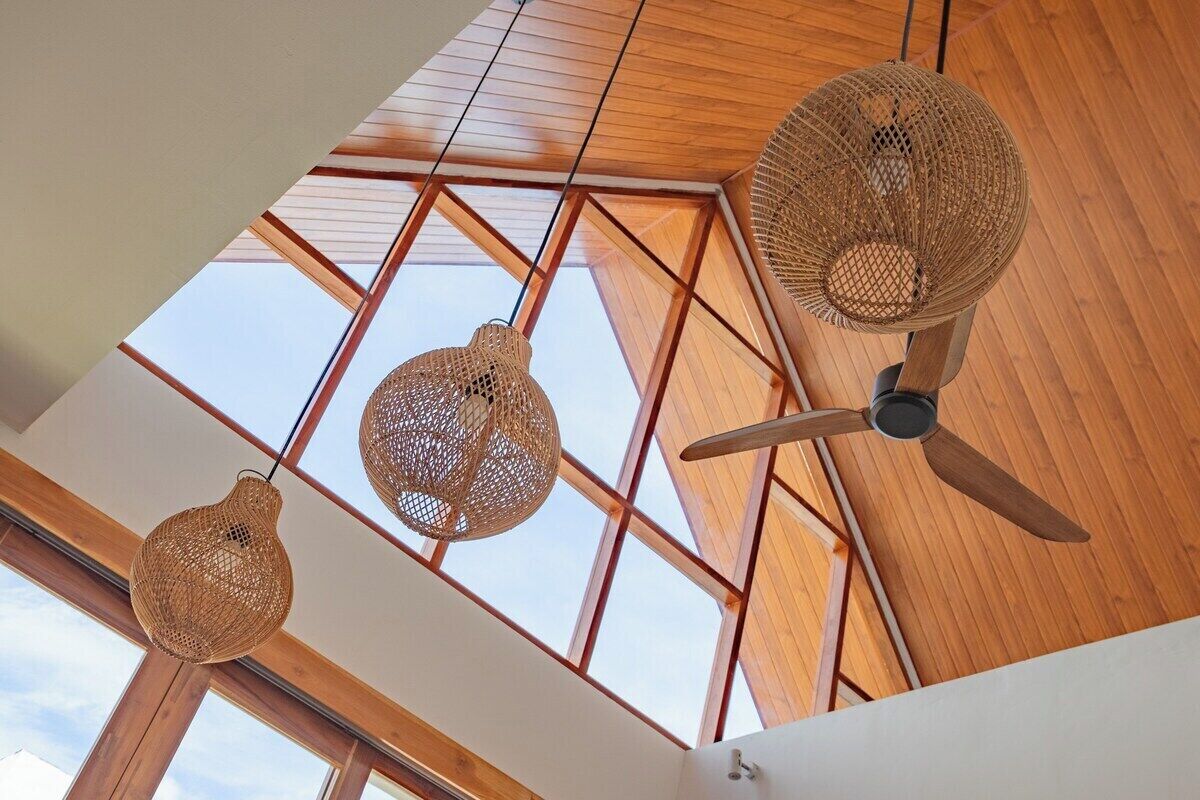 Ein Zimmer mit hoher Decke, Holzbalken und einem Oberlicht. Drei gewebte Pendelleuchten und ein Deckenventilator sind vorhanden.
