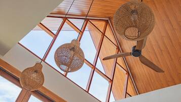Ein Zimmer mit hoher Decke, Holzbalken und einem Oberlicht. Drei gewebte Pendelleuchten und ein Deckenventilator sind vorhanden.