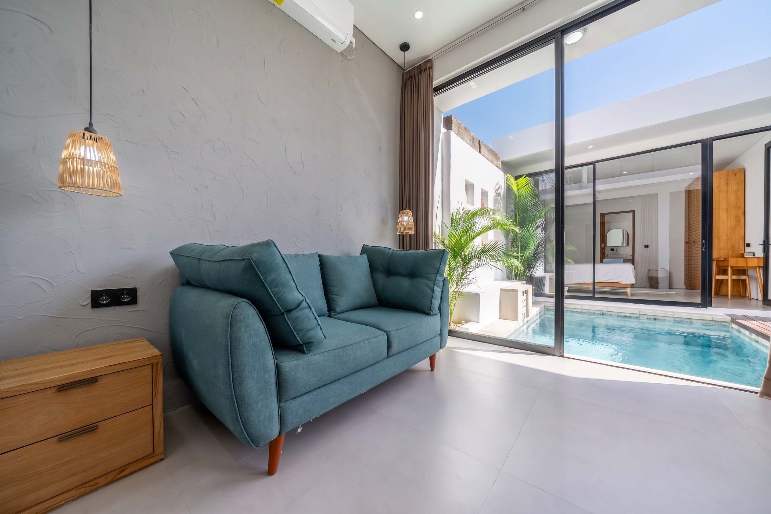 Una sala de estar moderna con un sofá color turquesa, una mesita de madera y un ventanal de vidrio que da a una zona de piscina.