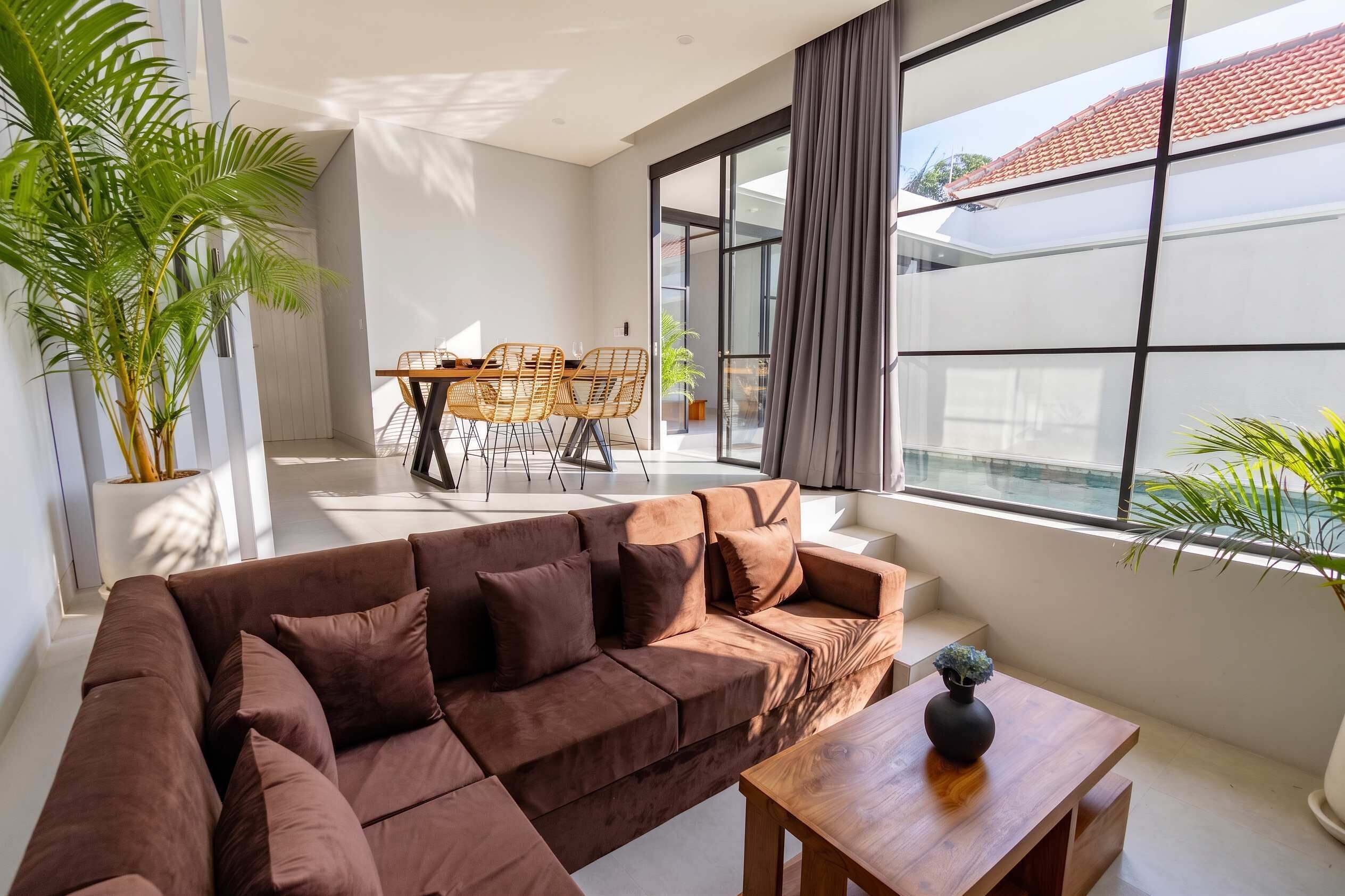 A modern living room with a sectional sofa, a dining table with chairs, and large windows offering a view of the outside.
