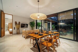 A modern dining area with a wooden table and chairs, a hanging light fixture, and a view through large glass doors. - 3BR Villa in Jungle Ubud • Private Pool & Patio (Gianyar Regency)