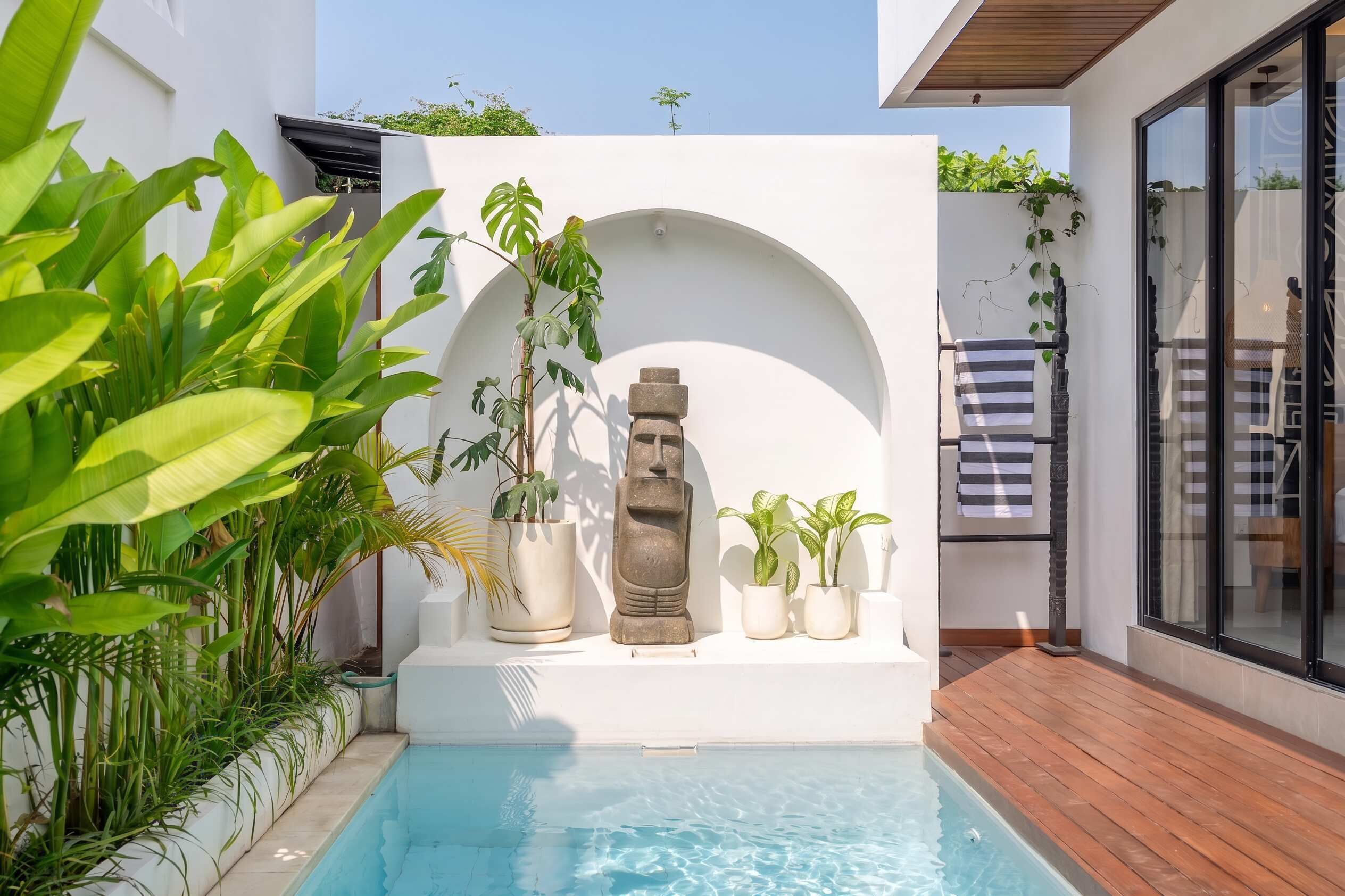 Ein moderner Poolbereich im Freien mit einer weißen Wand, Pflanzgefäßen und einer Steinskulptur.
