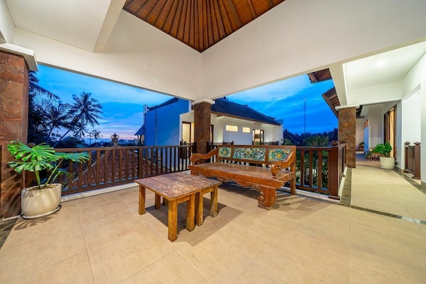 A spacious balcony with a wooden bench and table, potted plants, and a view of a house and palm trees. - Peaceful 1BR in Ubud w Pool, Patio & Lush Garden (Gianyar Regency)