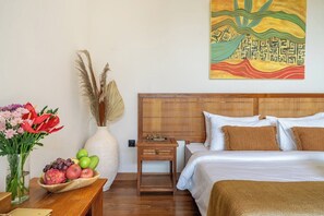 A hotel room with a wooden headboard, a bed with white bedding, a nightstand with a lamp, a vase with dried pampas grass, a bowl of fruit, and a painting on the wall. - Peaceful 1BR in Ubud w Pool, Patio & Lush Garden (Gianyar Regency)