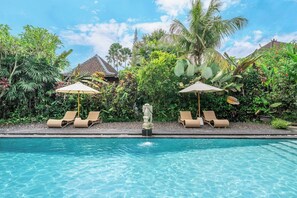 A swimming pool with lounge chairs and umbrellas, surrounded by lush greenery and a traditional building in the background. - Chic 1BR in Top Ubud's Spot w Spacious Pool+Garden (Ubud)