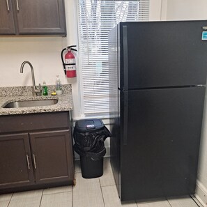 Una cocina con un lavamanos de acero inoxidable, un refrigerador negro, un cesto de basura y una ventana con persianas.