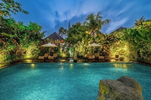 Ein Pool mit Liegestühlen und Sonnenschirmen, umgeben von üppiger Vegetation und blauem Himmel.