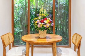 Een ronde houten tafel met een gevlochten mand waarin een kleurrijke bloemstelling staat, omringd door twee rieten stoelen met beklede zitplaatsen.