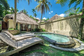 Uma área de piscina com espreguiçadeiras, uma estrutura com telhado de palha e um muro de pedra.