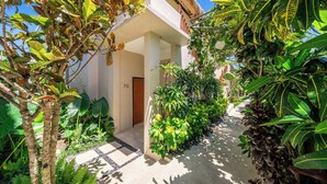 A tropical garden pathway leading to a building with a wooden door, surrounded by lush greenery and a clear sky.