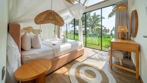 A bedroom with a large bed, a wooden nightstand, a round table, and a view of a green landscape through large windows. - New Tropical Villa+Large Pool+Perfect for Couples (Gianyar Regency)
